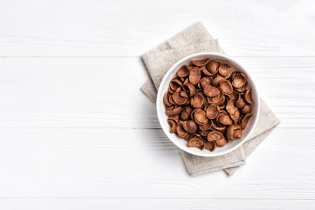 Top view of bowl with sweet chocolate cereal flakes for breakfast on white wooden background with...