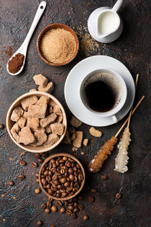 Top view of bowls with brown lump and granulated cane sugar, crystal sugar sticks, coffee beans, milk as ingredients for tasty drinks or beverages on dark concrete backgroundの写真素材
