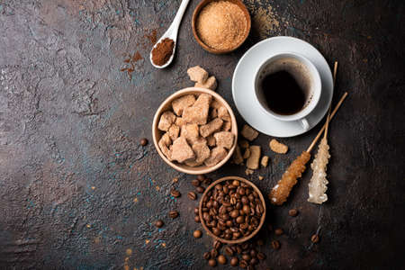 Top view of bowls with brown lump and granulated cane sugar, crystal sugar sticks, coffee beans, milk as ingredients for tasty drinks or beverages on dark concrete background with copy spaceの写真素材