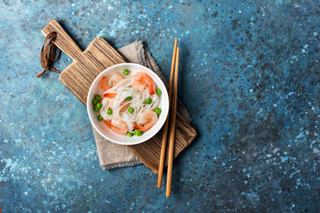 Top view of bowl with chinese rice noodles, tasty shrimps, green peas and wooden chopsticks on wood board and blue concrete backgroundの写真素材