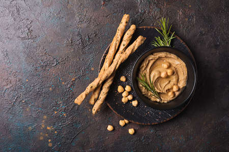 Top view of traditional homemade chickpea hummus snack with fresh green rosemary spice and grissini breadsticks in black bowl on dark concrete backgroundの写真素材