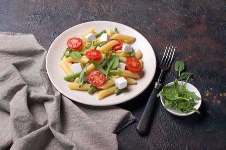 Tasty vegetarian penne pasta with cherry tomatoes, soft cheese feta, green beans, spinach and arugula leaves on brown concrete backgroundの写真素材