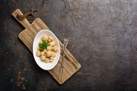 Bowl of small marinated or pickled champignon mushrooms with pepper, parsley and vintage silver fork on wooden board and brown concrete background with copy spaceの写真素材