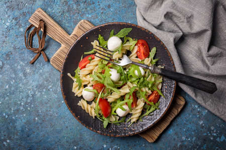 Top view of tasty vegetarian pasta with cherry tomatoes, soft cheese mozzarella and green arugula leaves on blue concrete backgroundの写真素材