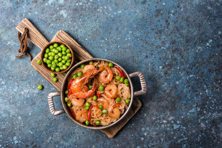 Top view of frying pan with chinese rice noodles, tasty prawns, green peas in bowl and red pepper on wooden board and blue concrete backgroundの写真素材