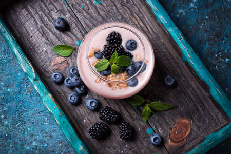 Top view of tasty yogurt dessert with blueberry, blackberry, oatmeal flakes and mint in portion glass on wooden tray and blue concrete backgroundの写真素材