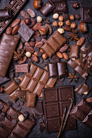 Pieces of broken tasty milk and dark chocolate bars with cocoa beans and hazelnut on black concrete backgroundの写真素材
