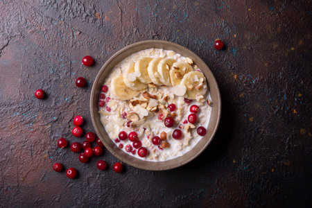 Top view of oatmeal with fresh cranberry, hazelnuts, almonds and banana slices for healthy breakfast on dark concrete backgroundの写真素材