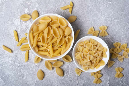 Top view on variety of italian raw pasta in white bowls on grey concrete background