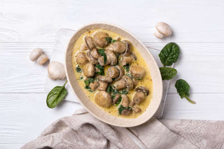Top view of champignon mushrooms in bowl with spinach leaves and cream sauce on white wooden backgroundの写真素材