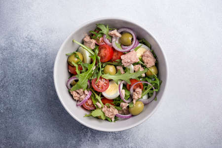 Top view of healthy and tasty salad with tuna fish, fresh arugula, cherry tomatoes, quail egg, onion and green olives on grey concrete backgroundの写真素材