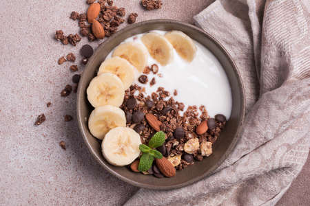 Top view of granola with yogurt, fresh banana, chocolate drops and almond nuts for healthy breakfast on brown concrete backgroundの写真素材