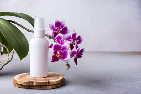 Mockup of skincare cosmetic product. White bottle of eco lotion on wooden pedestal with beautiful orchid flowers on modern grey backgroundの写真素材