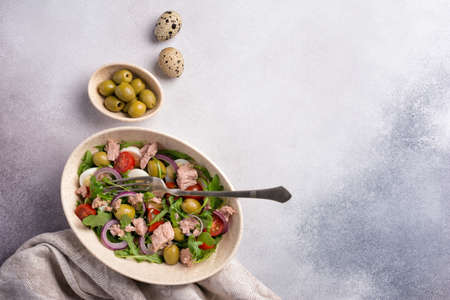 Top view of healthy and tasty salad with tuna fish, fresh arugula, cherry tomatoes, quail egg, onion and green olives on grey concrete backgroundの写真素材