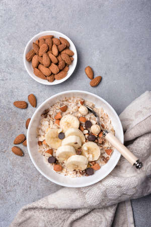 Top view of oatmeal with fresh banana slices, almond, hazelnut and chocolate drops for healthy breakfast on grey concrete backgroundの写真素材