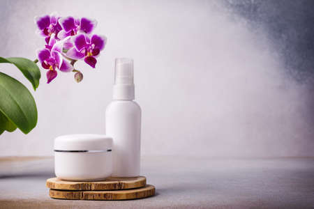 Mockups of skincare cosmetic products. White bottle of eco lotion and jar of cream on wooden pedestal with beautiful purple orchid flowers on modern grey backgroundの写真素材