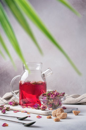 Beautiful flower pink sweet tea in glass teapot with dry rose buds and cane sugar on grey kitchen backgroundの写真素材