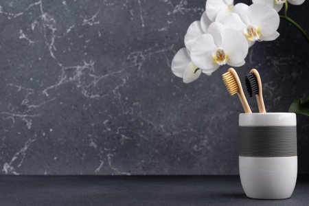 Eco friendly bathroom concept. Couple of bamboo toothbrush in a glass with white orchid flowers on dark grey background with copy spaceの写真素材