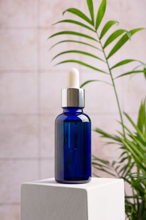 Mockup of skincare cosmetic product. Blue bottle of eco or natural serum on white cube pedestal with green plant on bathroom backgroundの写真素材