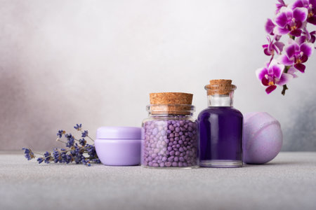 Spa wellness set with lavender flowers, orchid, essential oil bottles, cream jar, and purple granules for aromatherapy and natural skincare on a grey textured background.の写真素材