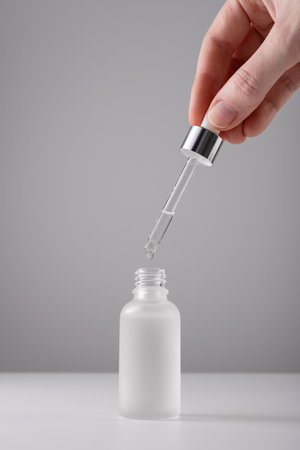 Human hand holding serum bottle with pipette on gray background. Close-up frosted glass container for skin care beauty product.の写真素材