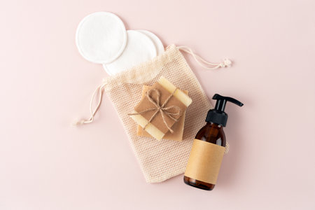 A collection of natural skincare essentials arranged on a gentle pink background. Features a pump bottle, handmade soap, and reusable cotton pads in a mesh pouch, promoting eco-friendly beauty.の写真素材