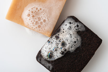 Closeup of two soap bars with foamy bubbles, one brown charcoal and one beige, on a white...