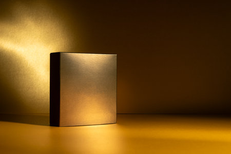Golden square box standing on a glowing golden surface with dramatic light and shadow. Elegant minimal packaging for cosmetic or luxury product branding mockup in studio setting.の写真素材