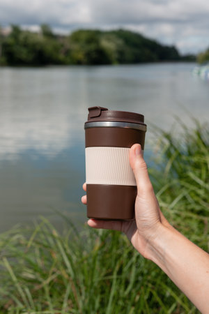 Hand holding a reusable thermo coffee cup near a peaceful river, surrounded by nature. Eco-friendly lifestyle, morning relaxation, outdoor leisure and sustainability concept.の写真素材