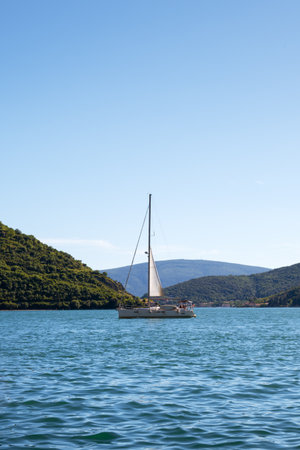 Sailboat drifting on calm blue water between green hills under a clear summer sky. Peaceful seascape...