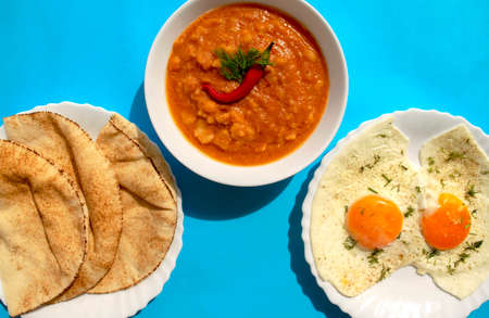 Famous traditional Arabic breakfast - peeled fava beans with chili pepper in a white bowl, pita bread, and fried eggs on a blue background. Top view. Foul Mudammas, Egyptian beans, flatbread, eggsの写真素材
