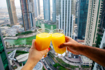 Female and male hands cheer glasses of fresh cold orange juice with city skyscrapers view of modern city Dubai. Two glasses of orange juice on cityscape background.の写真素材