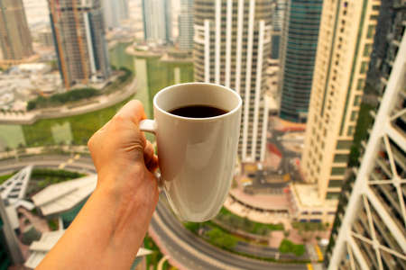 Female hand holds big white cup of morning coffee with city skyscrapers view of modern city Dubai. Coffee cup on city background.の写真素材