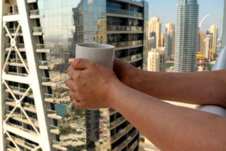 Middle age female holds big white cup of coffee with city skyscrapers view of Dubai Marina. Coffee cup on city background.の写真素材