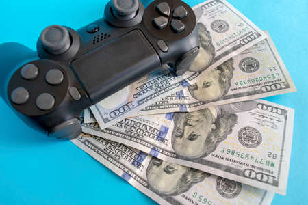 Black game joystick with us dollars on blue background. Playing for money concept. Game pad with money.の写真素材
