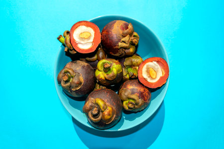 Mangosteen fruit in turquoise plate on blue background, top view. Many fresh purple mangosteens with one opened on the top of pile.の写真素材