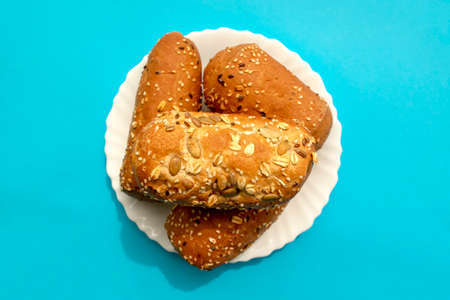 Multigrain mini bread in white plate on blue background. Flat lay, top view. Brown bread with different seeds - chia, pumpkin, flax, sesame. Small loafs.の写真素材