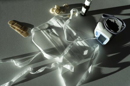On a light background with bright lighting and contrasting shadows are a white disposable protective medical mask made of non-woven material, plasters, fingertips and a tonometer. Medical concept.の写真素材
