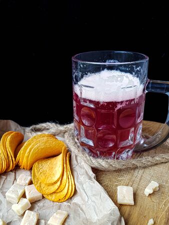 A glass of beer with foam on a dark background with a snack of chips and cheese on craft paper and a wooden kitchen Board. High quality photoの写真素材