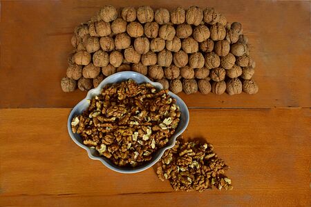 A bowl with walnut kernels and many walnuts folded in the form of a prism on a wooden table.