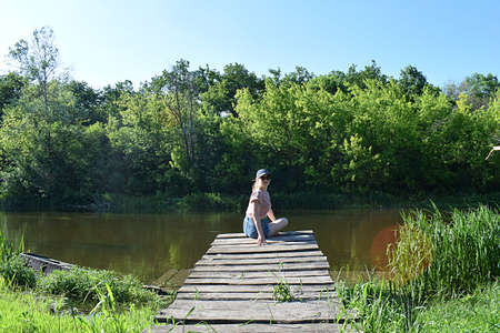 A girl sits on a wooden pier on the river bank on a summer sunny day.の写真素材