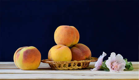 Ripe peaches in a wicker basket and a mallow flower on a wooden table.