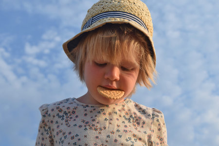 Little girl holding a cookie in her teeth. Portrait against the sky.の写真素材