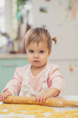 Adorable baby in an apron is rolling out the dough in the kitchenの写真素材