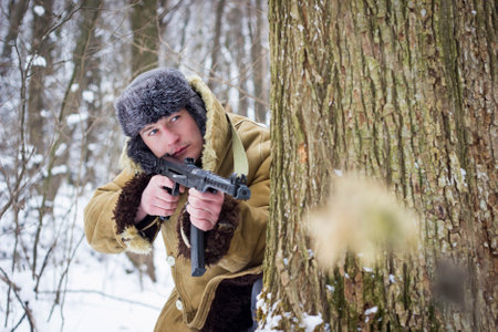 soldier in old coat and fur hat standing with a gun in the winter forest