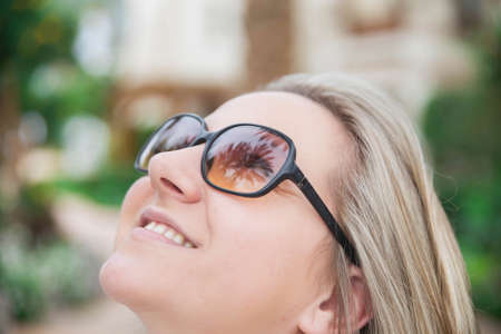 attractive woman in sunglasses smiling looking up in the garden and in her glasses palm trees are reflectedの写真素材