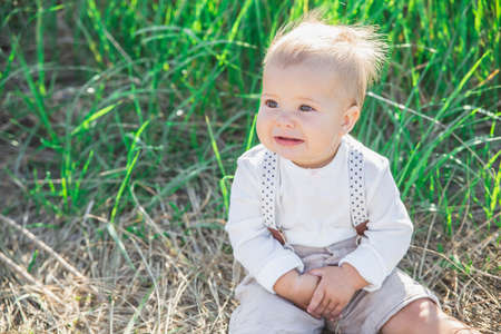 beautiful blond baby in a suit with suspenders sits in the grassの写真素材