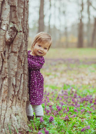 blond baby in a colored dress peeps out from behind a tree in the forestの写真素材