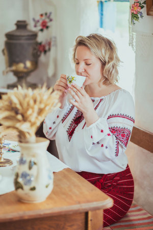Beautiful peasant woman in embroidered clothes drinks tea in an old Ukrainian hutの写真素材