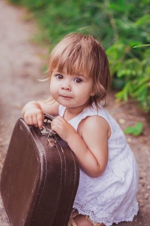Charming baby in a white sundress tries to open an old suitcaseの写真素材